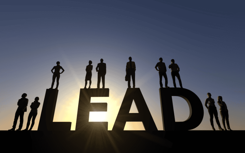 Silhouetted businesspeople standing on giant letters spelling "LEAD" at sunrise, symbolizing leadership, guidance, and influence in a corporate or professional setting.